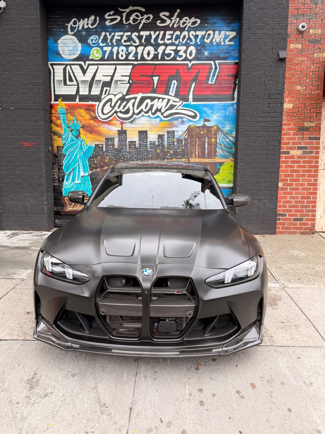 Satin black BMW M4 in front of the Lyfestyle Customz mural in Queens